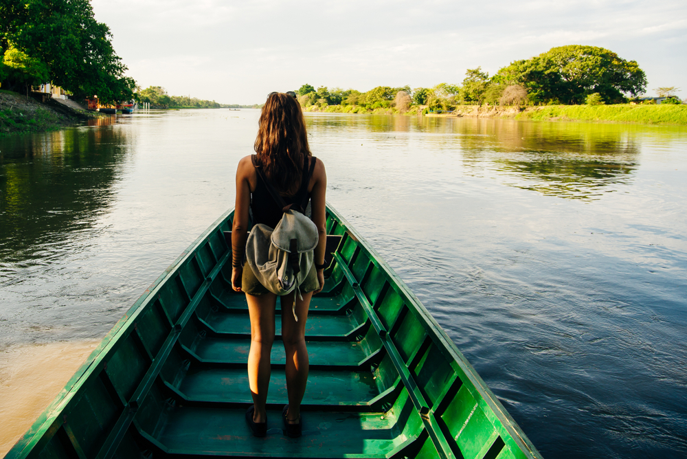 Woman with backpack at the bow of a long green boat on a wide river lined with tropical trees at golden hour